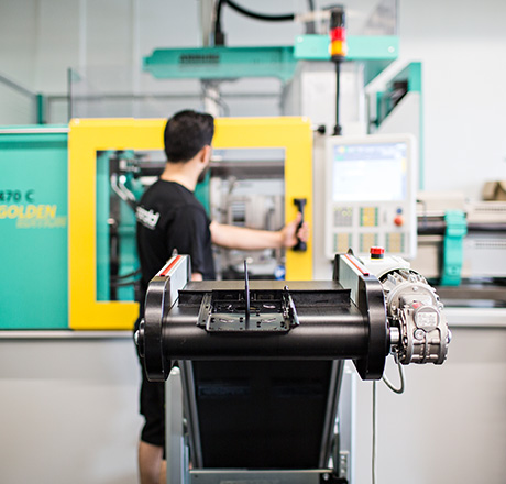 Im Vordergrund eine Maschine, im Hintergrund ein Mitarbeiter mit der Hand am Griff einer anderen Maschine Im Vordergrund eine Maschine, im Hintergrund ein Mitarbeiter mit der Hand am Griff einer anderen Maschine